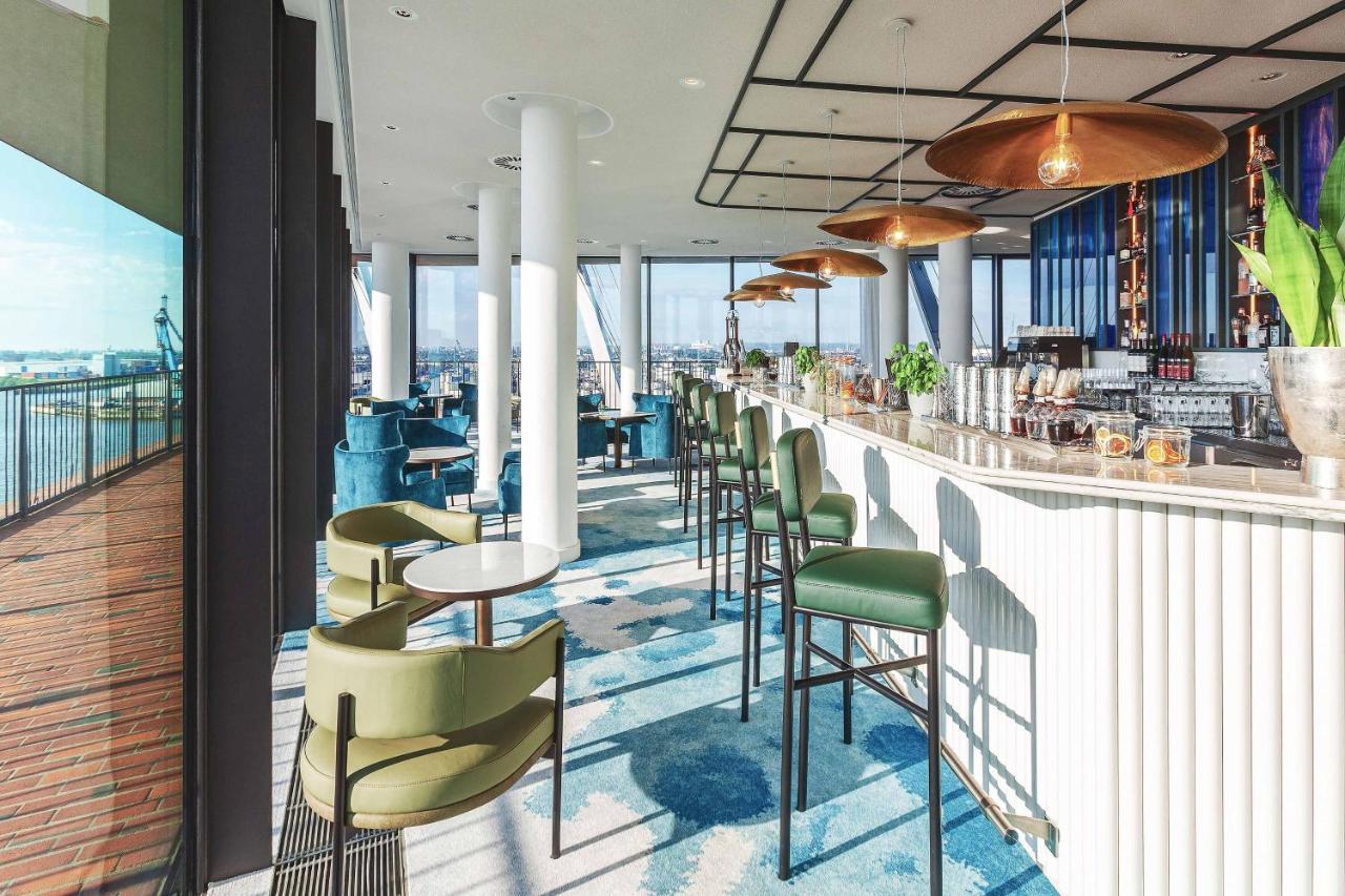 The Westin Hamburg Elbphilharmonie Hamburgo Exterior foto The photo depicts a modern bar or lounge area with a stylish design. There are high-backed green chairs surrounding small circular tables, arranged in an open space that has large windows showcasing a view of the outside. The bar area is sleek, featu